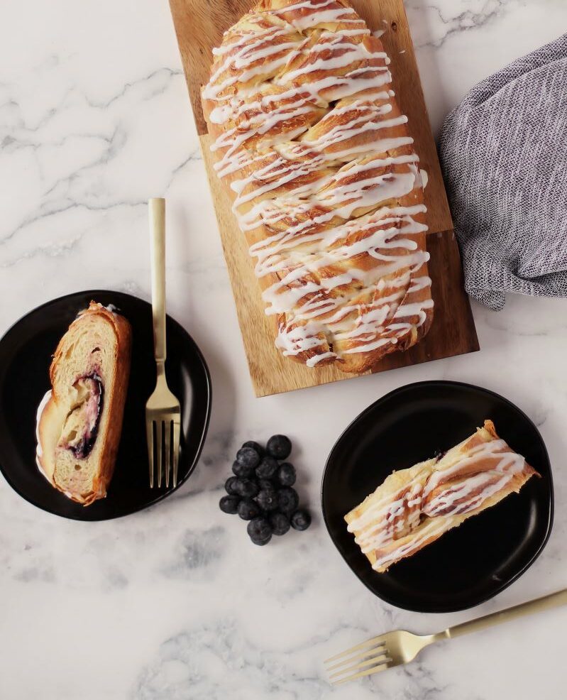 Blueberry & Cream Cheese pastry on cutting board with two slices of pastry on plates next to it.