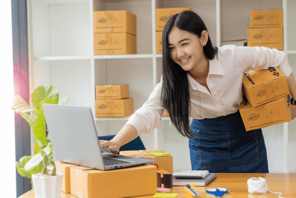 Young woman, holding two boxes and typing on a computer. More boxes on shelves behind her. Boxes are all branded with Butter Braid Pastry information. She is a volunteer for a group fundraiser.