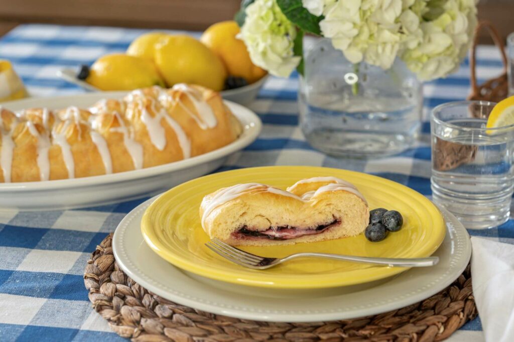 A slice of a Blueberry & Cream Cheese pastry is on a plate. A full, baked pastry is on a platter in the background. It is all set on a spring-themed table.