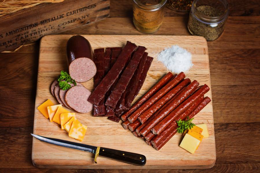 A wood cutting board with meat sticks, jerky, summer sausage, and cheeses.